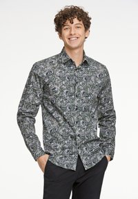 Langsleeve shirt met een groen, grijs en zwart abstract bloempatroon, met knopen en een kraag. Zwarte broek wordt gedragen.