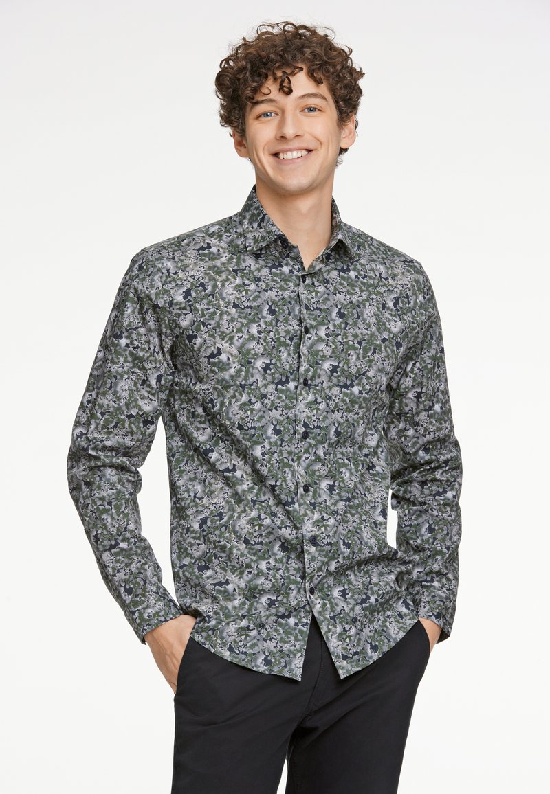 Langsleeve shirt met een groen, grijs en zwart abstract bloempatroon, met knopen en een kraag. Zwarte broek wordt gedragen.