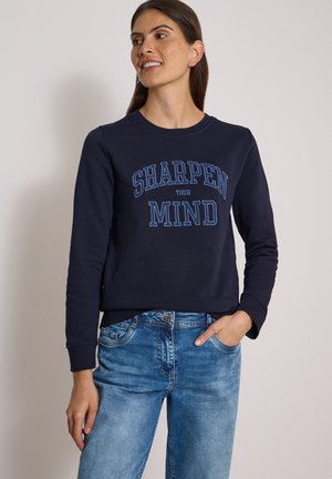 Frau trägt einen dunkelblauen Sweatshirt mit dem Text "SHARPEN YOUR MIND" und blaue Jeans, lächelnd mit einer Hand in der Tasche, steht vor einem einfarbigen Hintergrund.