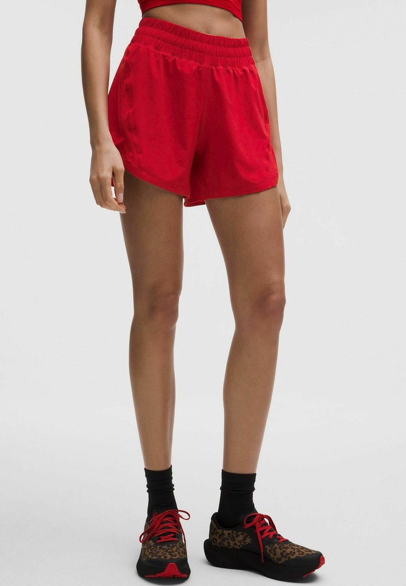 Žena u crvenim sportskim šortsevima, crnim čarapama i tenisicama za trčanje s leopardovim uzorkom i crvenim vezicama, stoji ispred jednostavne svijetle pozadine.