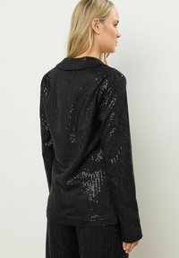 Donna bionda vista di spalle che indossa un blazer nero con paillettes e pantaloni abbinati, in piedi contro un muro chiaro e semplice.