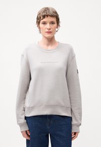Lys grå genser med rund hals, ribbestrikkede mansjetter og nederkant. Har et tekstgrafikk foran og et lite logomerke på den ene ermen.
