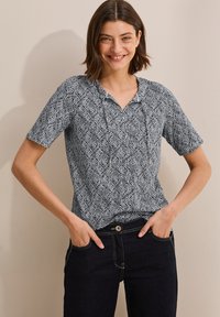Kortärmad blus med V-hals, prydd med ett svartvitt lövmönster på mjuk tyg; matchad med mörka jeans och synliga fickdetaljer.