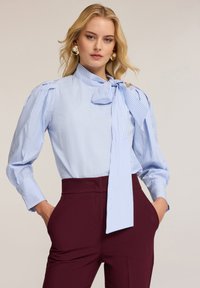 Hellbluse mit Streifen, einer Schleife, puffigen Ärmeln und Knopfmanschetten, kombiniert mit hochgeschnittenen kastanienbraunen Hosen mit Taschen.