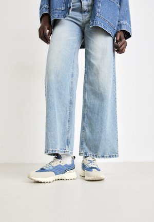 Persoon die lichtblauwe wijdbenige jeans, een spijkerjasje, witte sokken en blauwe en witte sneakers draagt, staand op een lichte vloer.