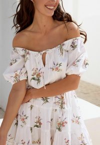 Witte off-the-shoulder jurk met korte puffmouwen, voorzien van een bloemenpatroon in roze, paars en groen, en een strik bij de hals.