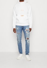 Sudadera blanca con capucha y un pequeño logo naranja en el frente, combinada con jeans desgastados de color azul claro y zapatillas blancas.