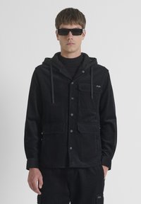Chaqueta de pana negra con capucha, que cuenta con botones a presión, dos bolsillos frontales y un logo bordado en el pecho. Lleva puesta una camiseta de cuello alto negra.