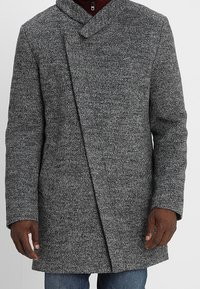 Grijze, gestructureerde overjas met een sjawlboord, asymmetrische sluiting aan de voorkant en zijzakken, gedragen over een maroon shirt.