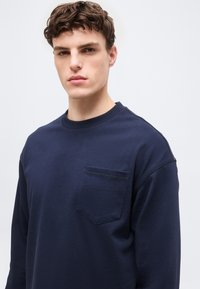 Νέος άνδρας με κοντά σγουρά μαλλιά που φοράει μπλε σκούρο πουκάμισο με μακριά μανίκια και τσέπη στο στήθος, κοιτάζοντας ελαφρώς προς το πλάι.