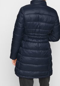 Tummansininen untuvatakki, jossa on vaakasuorat saumat, korkea kaulus ja vyötä liioitellusti. Valmistettu kiiltävästä, sileästä materiaalista ja siinä on yksivärinen muotoilu.