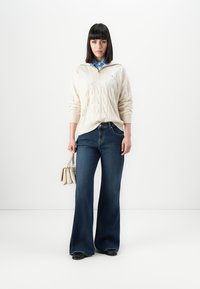 Femme aux cheveux noirs raides portant un pull crème en tricot torsadé sur une chemise bleue, un jean évasé foncé, des chaussures noires et tenant un sac à main beige.