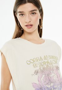 Camiseta de algodón crema de corte holgado con mangas enrolladas, que presenta un diseño gráfico de serpiente y flores en colores púrpura y dorado. Textura suave, estilo casual.