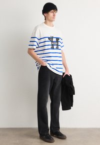 Gestreiftes Baumwoll-T-Shirt in Weiß und Blau mit Logo, kombiniert mit schwarzen Cordhosen. Die Person trägt eine schwarze Mütze und hält eine Jacke.