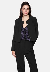 Black cropped jacket with round neckline and button closure. Worn over a purple patterned blouse. Trousers are also black with pleats.