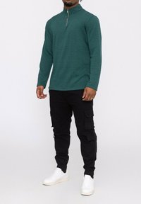 Haut à manches longues zippé vert avec un motif texturé, associé à un pantalon cargo noir avec de multiples poches et des baskets blanches.