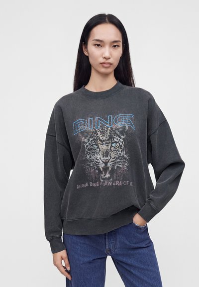 Femme aux longs cheveux noirs portant un sweat-shirt noir délavé avec un motif léopard et le mot Bing, associé à un jean bleu.