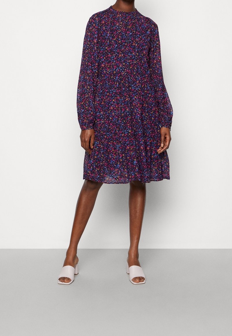 Femme portant une robe longue manches longues, longueur genou, de couleur violet foncé avec un petit motif floral multicolore et des chaussures blanches ouvertes à l'avant à talons blocs.