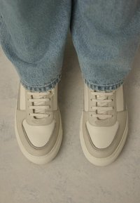 Baskets en cuir beige avec accents gris et semelles en caoutchouc. Elles comportent des lacets plats et un bout arrondi. Portées avec un jean bleu clair retroussé.