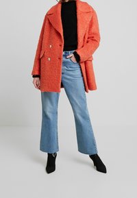 Femme portant un manteau orange surdimensionné en texture, un pull noir, un jean clair à jambes larges et des bottines à talons noires, se tenant devant un fond uni.
