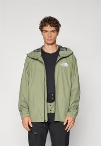 Leichte, olivgrüne Jacke mit Kapuze, elastischen Bündchen und einem schwarzen Innenfutter. Mit einem Logo auf der linken Brust und mit Reißverschlusstaschen.