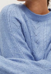 Svetle modrý svetr s pleteným vzorem a texturou, který má na sobě osoba, zobrazuje detaily horní části trupu a krku.