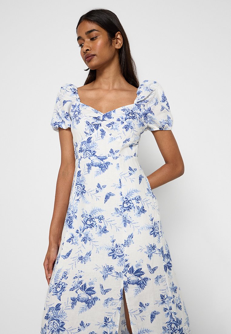 Weißes Kleid mit blauem Blumenmuster, Puffärmeln und quadratischem Ausschnitt. Verfügt über einen Schlitz vorne für mehr Bewegungsfreiheit.