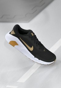Sort og guld Nike løbesko med hvid sål, snørebåndene bundet, præsenteret på en glat, lys grå overflade med geometrisk refleksion fra loftet.