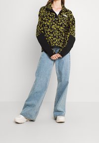 Pullover in pile a stampa leopardata nera e verde con colletto zip, abbinato a jeans a gamba larga azzurri e sneakers bianche.