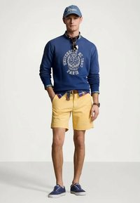 Άνδρας φορώντας μπλε καπέλο, ναυτικό φούτερ με λογότυπο πανεπιστημίου, καρό πουκάμισο, κίτρινα σορτς, μπλε αθλητικά παπούτσια και γυαλιά ηλίου κρεμασμένα από το γιακά του φούτερ.