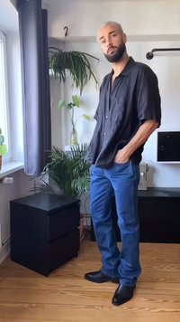 Homem com cabelo loiro curto e barba está em um ambiente fechado, vestindo uma camisa preta, jeans azuis e sapatos pretos, perto de plantas verdes e um armário preto.
