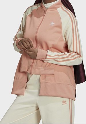 Person iført en pink og cremefarvet Adidas træningsdragt med lynlås foran, tre stribede ærmer og matchende bukser, der justerer sine ærmer.