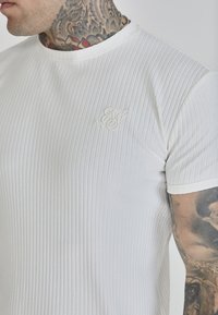 Tricou scurt alb cu mânecă scurtă, cu decolteu rotund și logo embosat pe piept. Dispune de un material texturat și o bordură contrastantă.