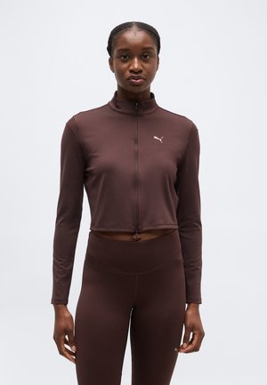 Femme portant un crop top à manches longues marron foncé avec fermeture éclair et un legging assorti taille haute, debout devant un fond clair uni.