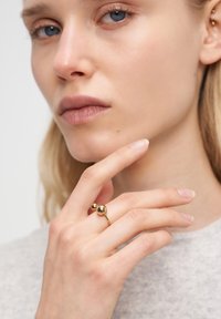 Guldring med två sfäriska accenter på en smal ring. Modellens hand vilar nära ansiktet, vilket framhäver ringens släta yta mot en ljusgrå bakgrund.