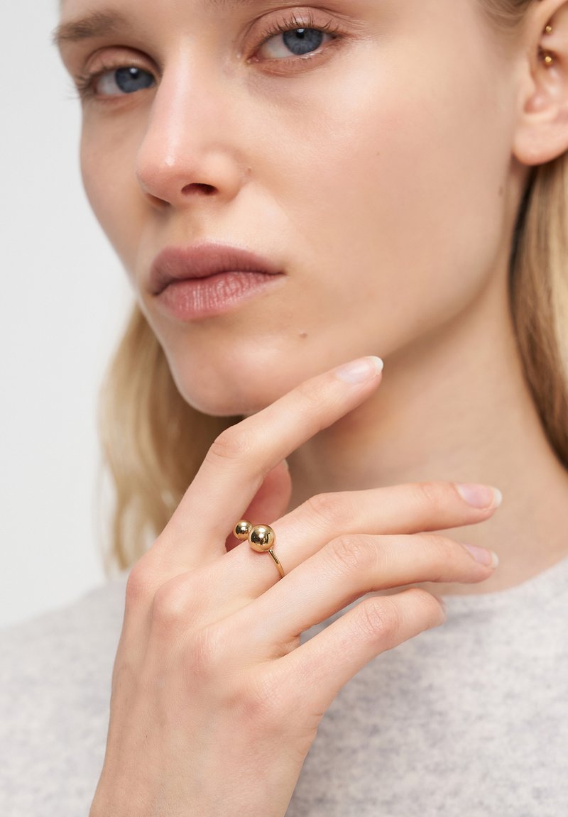 Guldring med två sfäriska accenter på en smal ring. Modellens hand vilar nära ansiktet, vilket framhäver ringens släta yta mot en ljusgrå bakgrund.