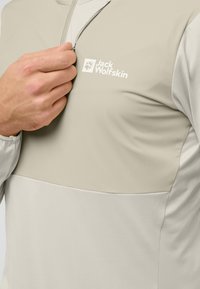 Lichtgewicht, beige hoodie met een ritskraag, voorzien van een donkerder beige paneel en het Jack Wolfskin-logo op de borst. Gladde textuur.