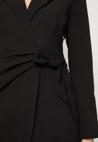 Vestido negro envolvente con mangas largas, que presenta un detalle de cintura anudada, tejido suave y solapas estructuradas. El diseño enfatiza una silueta ajustada.
