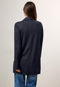 Blazer lungo nero con colletto a profilo e dettagli sartoriali, realizzato in un tessuto morbido e leggero, abbinato a jeans blu.