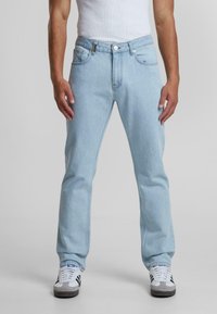 Jeans de mezclilla azul claro con un diseño de pierna recta, cinco bolsillos y cierre de botón. Combinados con zapatillas blancas con detalles en negro.