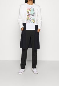 Lungo cappotto bianco con accenti blu navy, indossato sopra una maglietta a fumetti colorata. Pantaloni neri e sneaker bianche completano l'outfit.