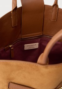 Bruine leren handtas met een gladde buitenkant en suède accenten. Het interieur heeft een bordeauxrode voering en een ritsvak. Het etiket vermeldt "Coccinelle, Authentiek Italiaans Leer."