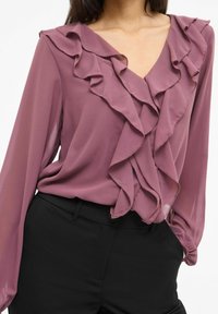 Lavendelbluse med volangdetaljer ved halsen og ermene, laget av lett stoff; kombinert med svarte, tettsittende bukser.