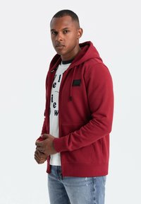 Sudadera roja con cremallera y bolsillo frontal, con un acento negro en el pecho, llevada sobre una camiseta gráfica blanca y jeans azul claro.
