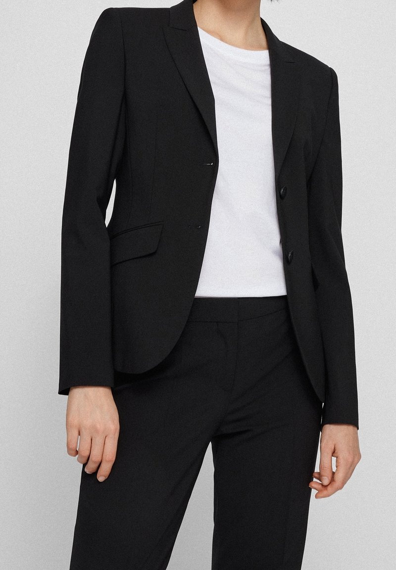 Svart skräddarsydd kavaj med två knappar, slagkragar, sidofickor och slät textur. Bärs med en vit t-shirt under.