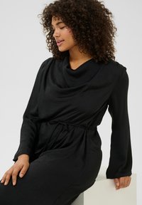 Vestido negro de manga larga con escote drapeado y cintura ajustable con cordón. Presenta una tela suave y texturizada, y una Silueta relajada.
