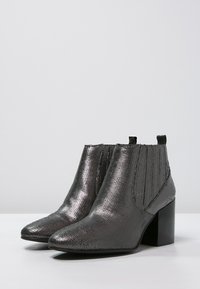 Metallisch silberne Knöchelstiefel mit spitzer Zehe, strukturierten Oberflächen, elastischen Seitenteilen und einem klobigen schwarzen Absatz.