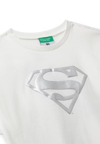 Bílý tričko s potiskem stříbrného loga Supermana na hrudi a zelenou visačkou United Colors of Benetton DC uvnitř límce.