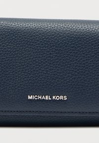 Πορτοφόλι από δέρμα σε ναυτικό μπλε χρώμα με υφή στην επιφάνεια και ορατές ραφές. Διαθέτει ασημί λογότυπο "Michael Kors" στο εμπρός μέρος.