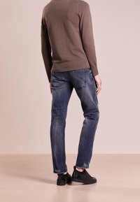 Suéter de punto marrón con puños acanalados, combinado con unos vaqueros de mezclilla azul desvaído de corte clásico. Unos tenis negros perforados completan el look.